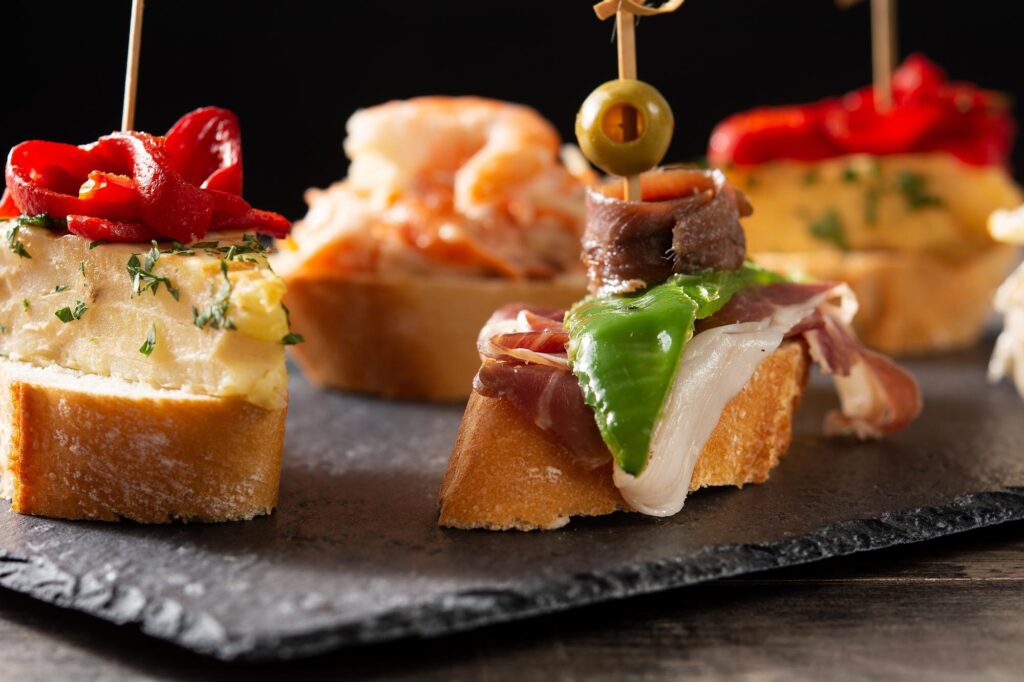 Imagen de un pincho de jamón, pimientoi, anchoa y una aceituna pinchada en un palo